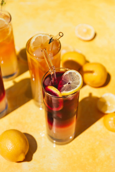 Hibiscus Mango iced tea blended with lemonade in a glass on a yellow table with full and sliced lemons in glass and on table