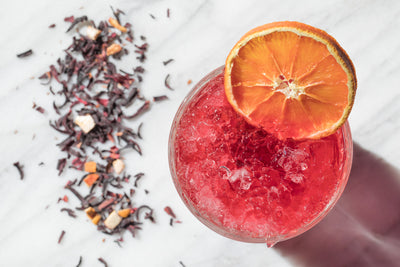 Glass of Hibiscus Crush with an orange garnish beside loose hibiscus tea.