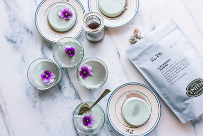 Several small glasses of light blue colored Blackberry Jasmine Panna Cotta with purple flower garnishes and a bag of iced tea on a marble counter.
