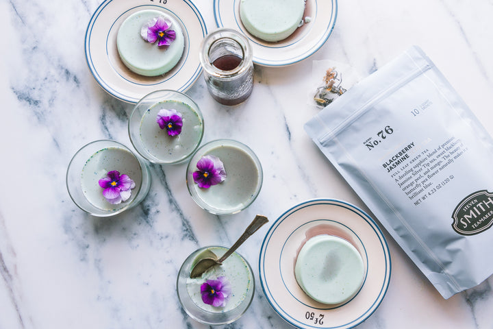Several small glasses of light blue colored Blackberry Jasmine Panna Cotta with purple flower garnishes and a bag of iced tea on a marble counter.