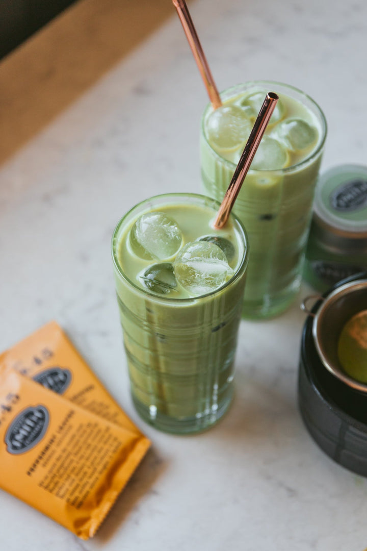 Two iced matcha glasses with straws and 2 peppermint leaves sachets on a marble table
