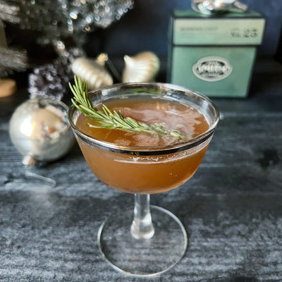 Coupe glass filled with Dash Away All cocktail with rosemary sprig garnish with Christmas ornaments and box of Morning Light Tea in background.