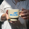 Person holding a mug with 'ART PERSON' text, wearing a white shirt.