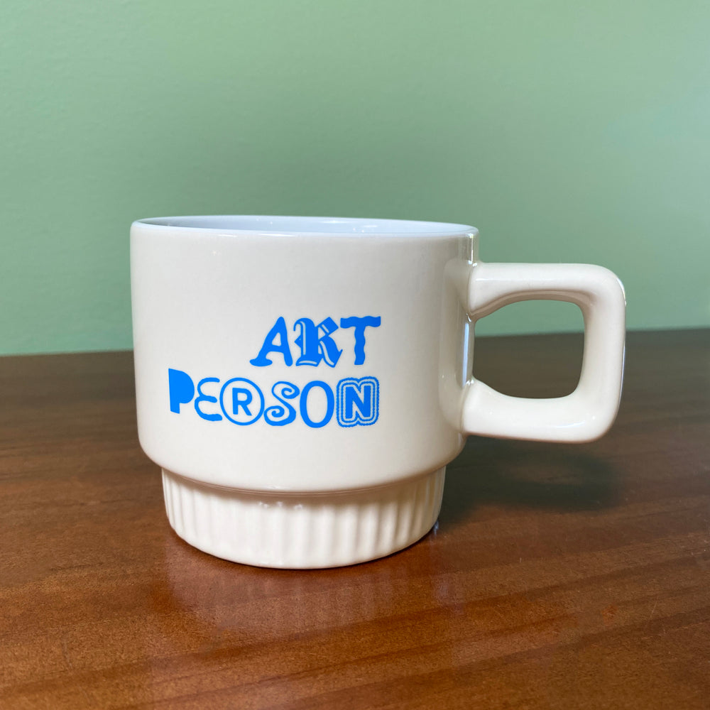 White mug with 'ART PERSON' text in blue on a wooden surface with a green background