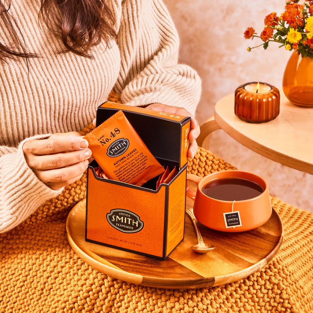 Person opening a box of Autumn Leaves to pull out a sachet alongside an orange teacup filled with tea.