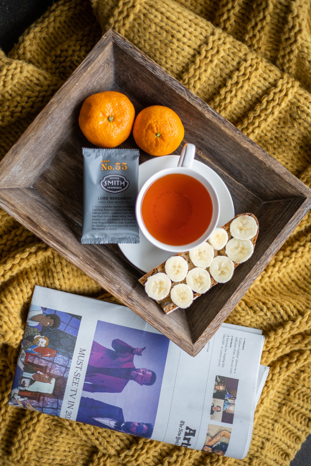 Tray with cup of black tea, a sachet of Lord Bergamot, a piece of banana toast and a newspaper.