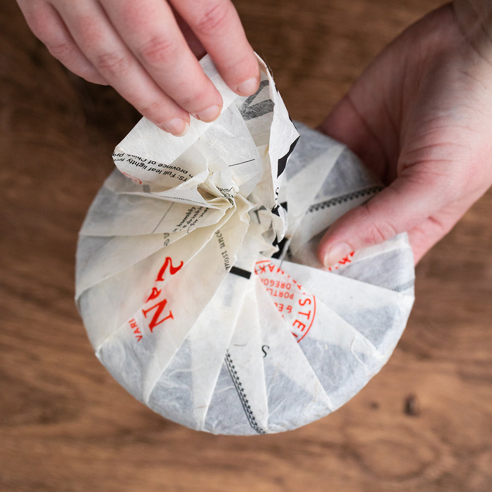 Hands unwrapping a pu-erh cake to show detail of the wrapping around the pu-erh.