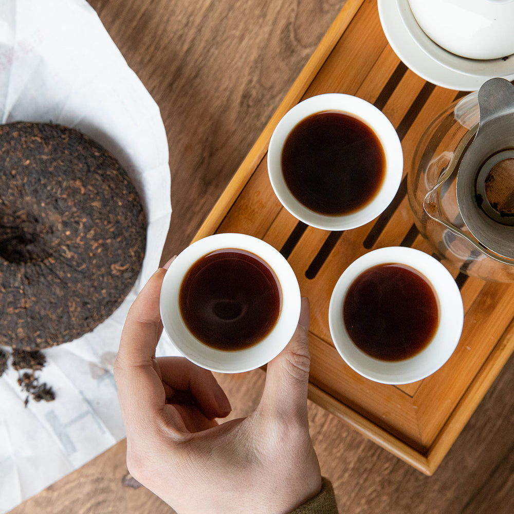 Hand reaching for a small gaiwan cup filled with brewed pu erh tea on a gaiwan tray.