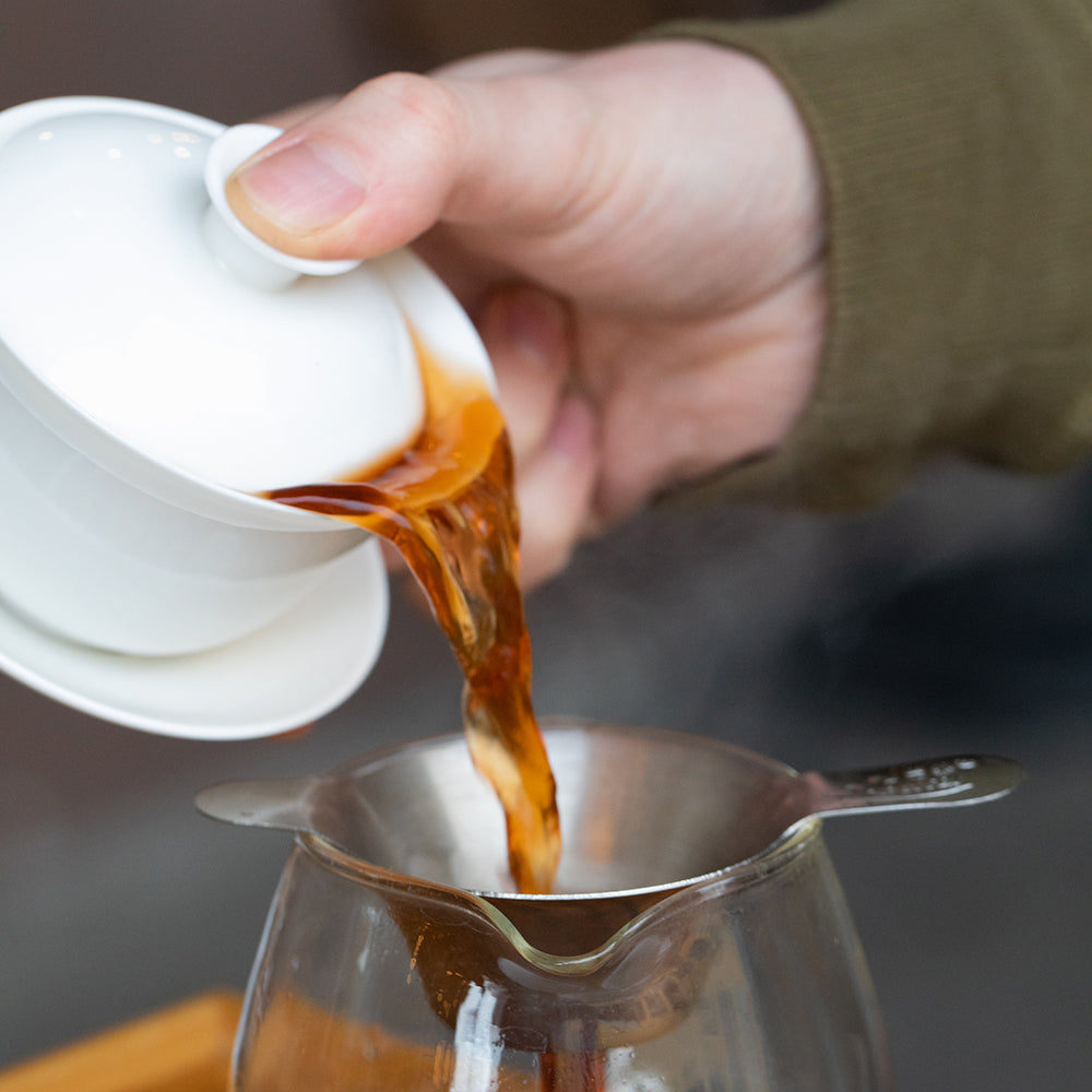 Hand holding gaiwan to pour tea over a strainer into a tea decanter.