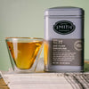 A silver tin of Steven Smith Teamaker Darjeeling No. 17 full leaf black tea stands next to a clear glass cup filled with amber tea, set on a stack of papers against a soft sage green background.