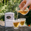 Brewed Double Roasted Oolong tea being poured from tea decanter into two glass gaiwan tea cups with a tin of Double Roasted Oolong in the background in an outdoor setting.