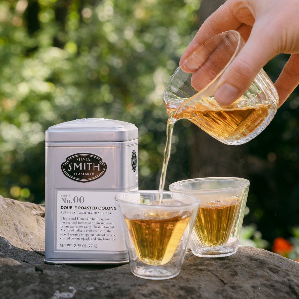 Brewed Double Roasted Oolong tea being poured from tea decanter into two glass gaiwan tea cups with a tin of Double Roasted Oolong in the background in an outdoor setting.