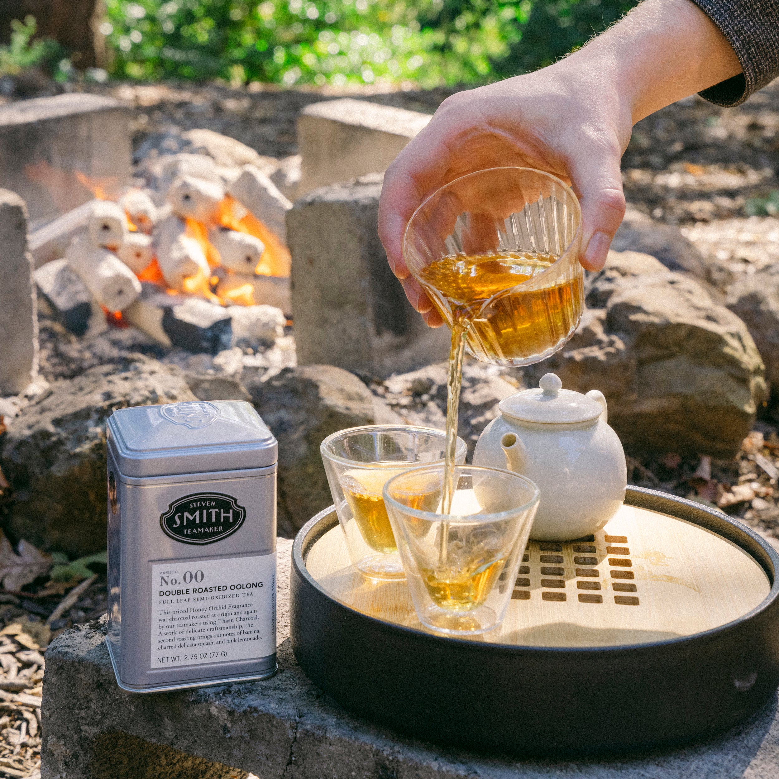 Brewed tea being poured from tea decanter into two glass tea cups with a tin of Double Roasted Oolong in the background in an outdoor setting.
