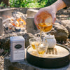 Brewed tea being poured from tea decanter into two glass tea cups with a tin of Double Roasted Oolong in the background in an outdoor setting.