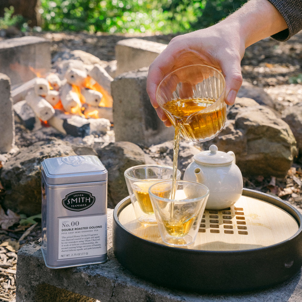 Brewed tea being poured from tea decanter into two glass tea cups with a tin of Double Roasted Oolong in the background in an outdoor setting.