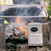 Tin of Double Roasted Oolong in the forefront on a rock with a camp fire and actively roasting tea in the background