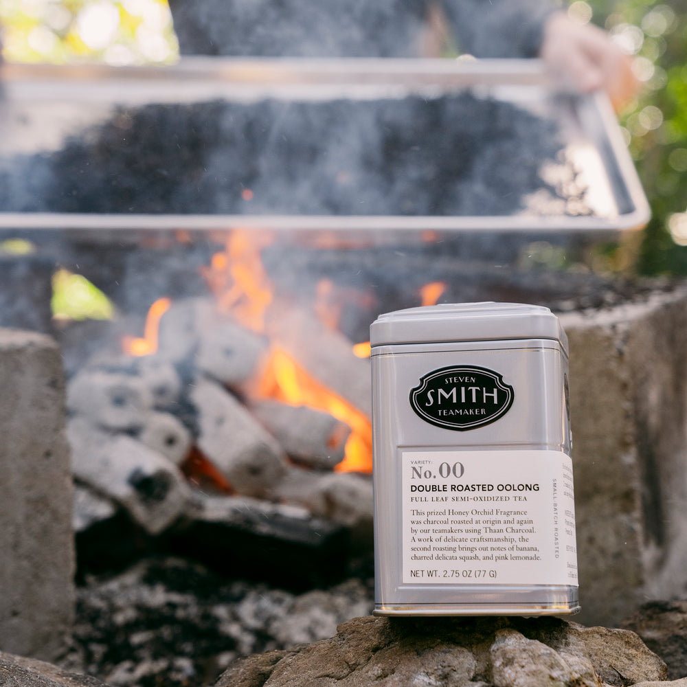 Tin of Double Roasted Oolong in the forefront on a rock with a camp fire and actively roasting tea in the background