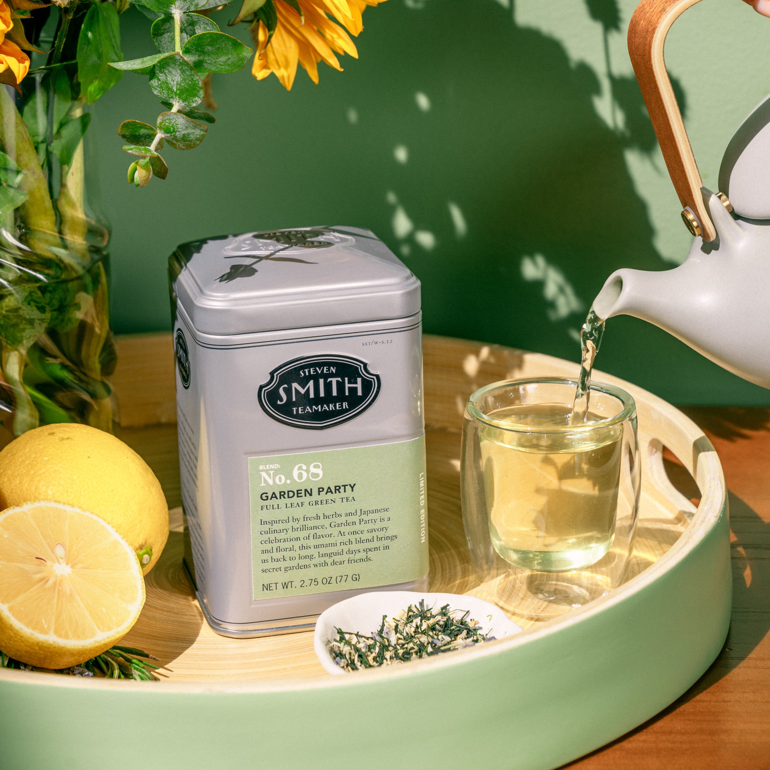 Grey tin of Garden party in a sunny setting surrounded by lemons, flowers, loose leaf tea and brewed tea being poured into a glass cup from a teacup.