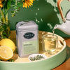 Grey tin of Garden party in a sunny setting surrounded by lemons, flowers, loose leaf tea and brewed tea being poured into a glass cup from a teacup.