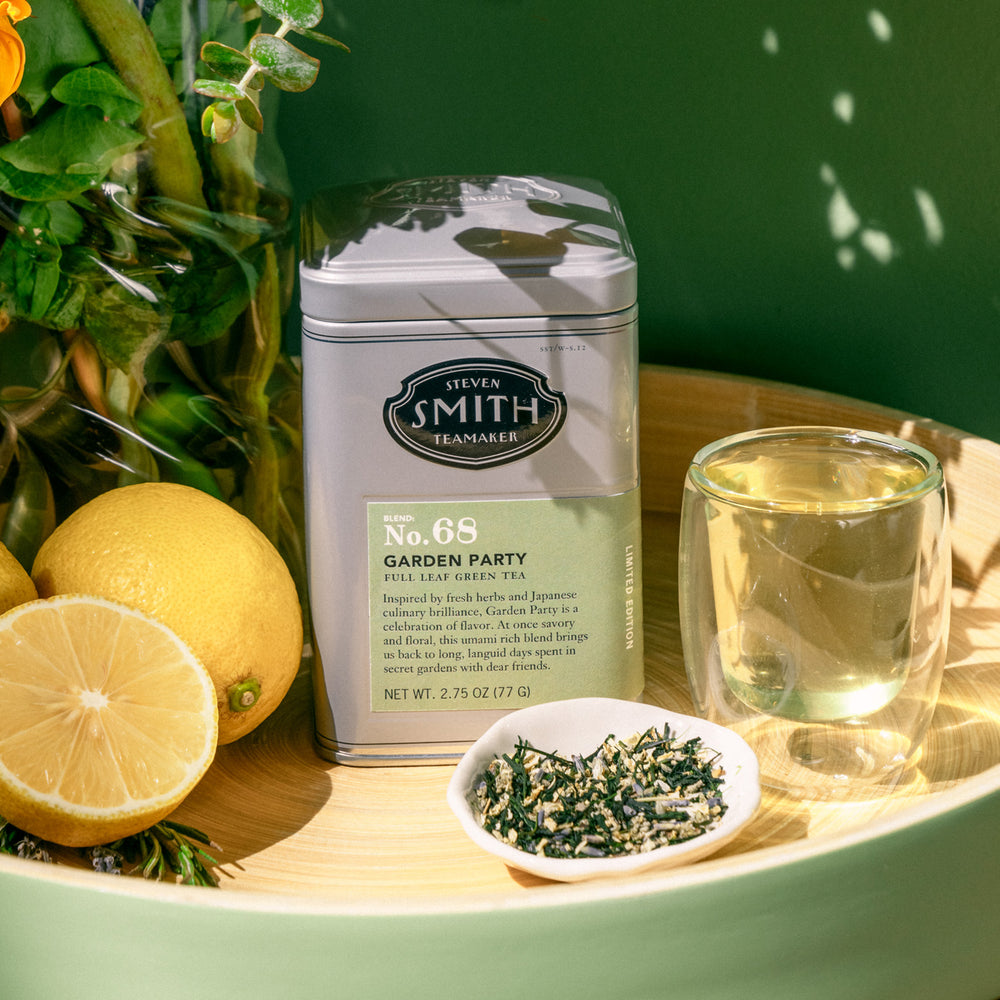 Grey tin of Garden party in a sunny setting surrounded by lemons, flowers, loose leaf tea and brewed tea.