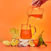 Person pouring tea into a glass mug with a ginger root and fruits on an orange background