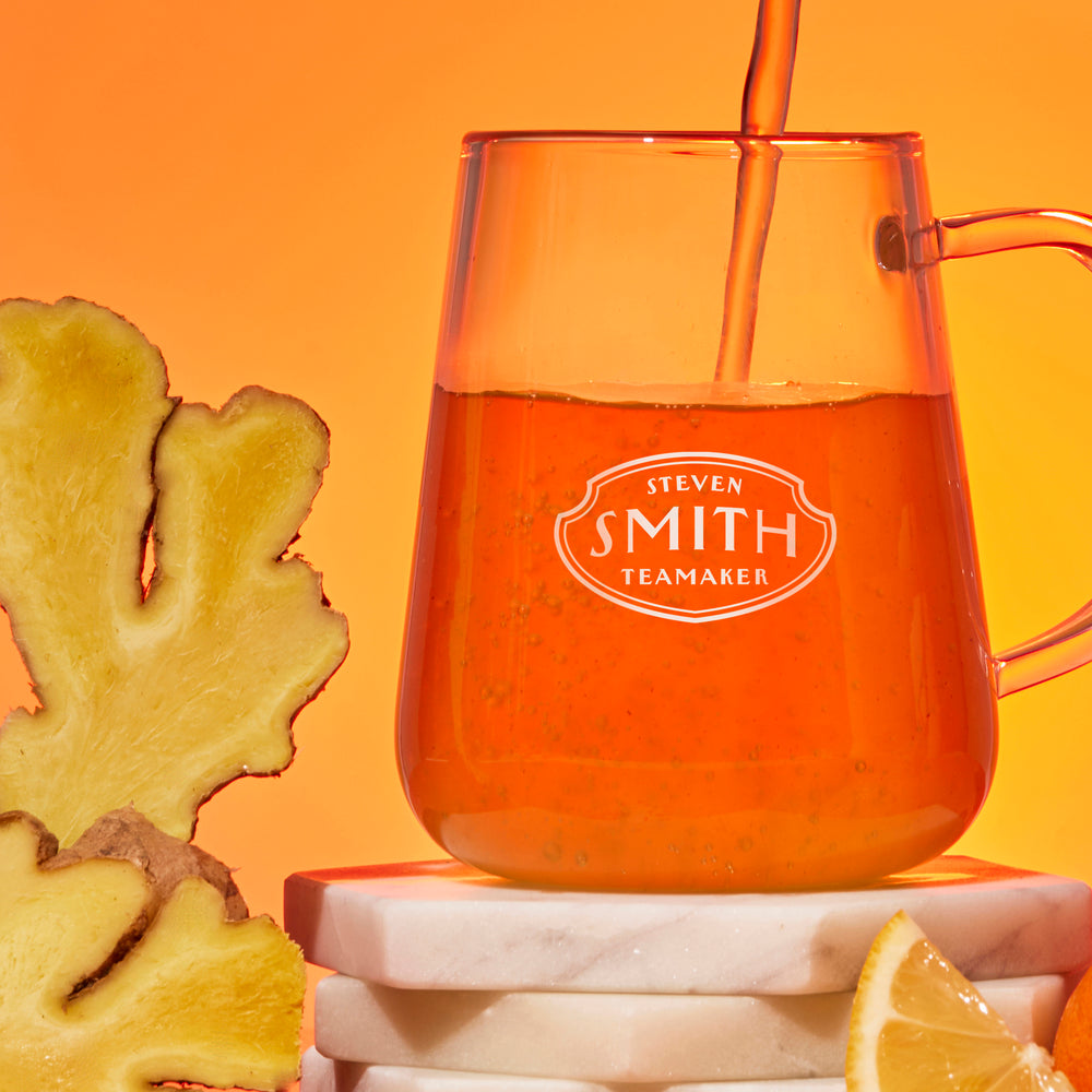 Glass mug with 'Steven Smith Teamaker' branding filled with tea, next to a piece of ginger and lemon on an orange background.