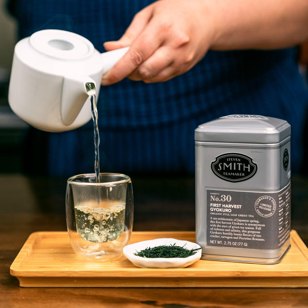 Tin of First Harvest Gyokuro on a wooden tray with a small bowl of loose tea leaves and a glass cup being filled with brewed tea.