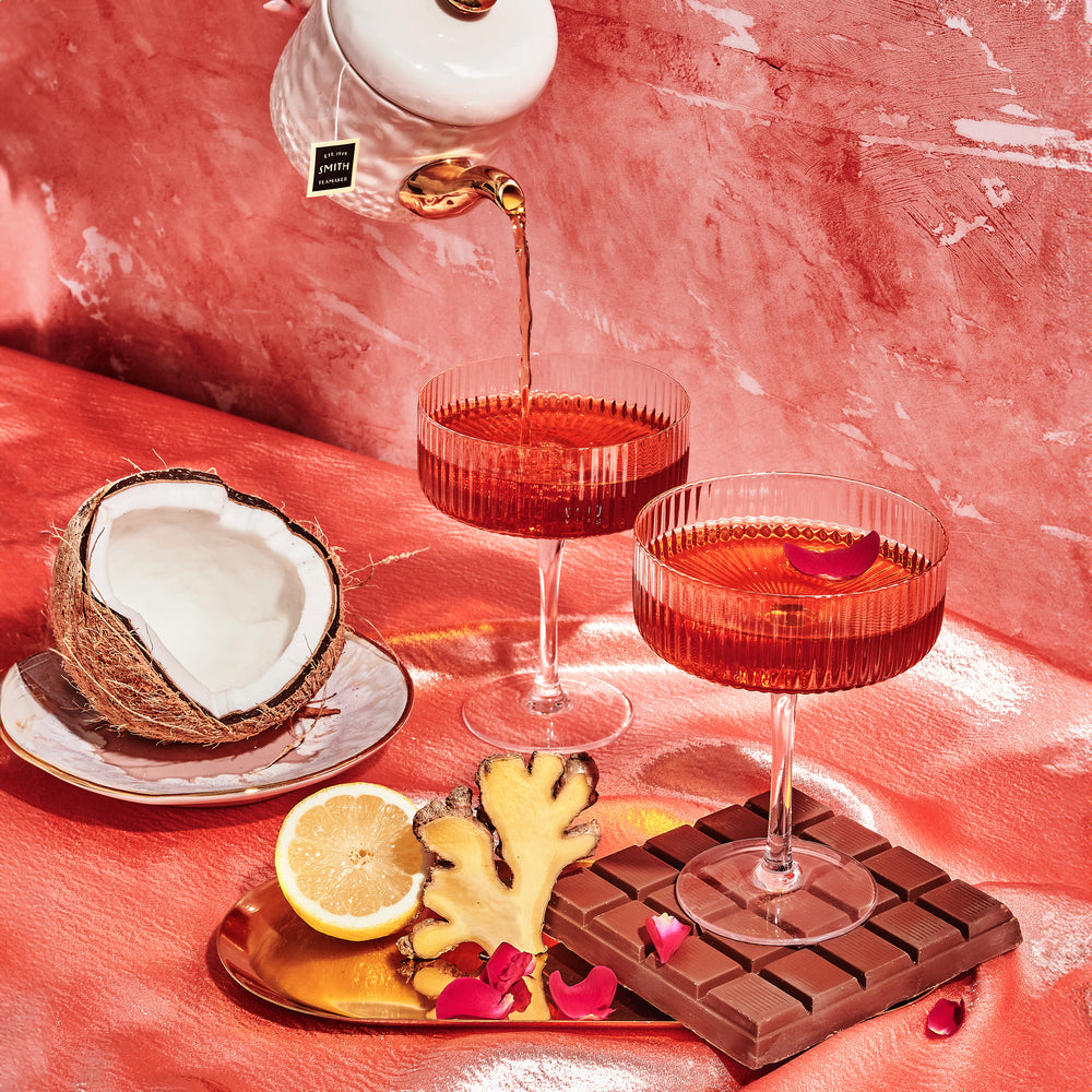 Two coup glasses filled with tea beside an opened coconut, a bar of chocolate, ginger and lemon.
