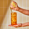 Hand squeezing juicy lemon into full glass of Lemon Black iced tea against a yellow tile background.