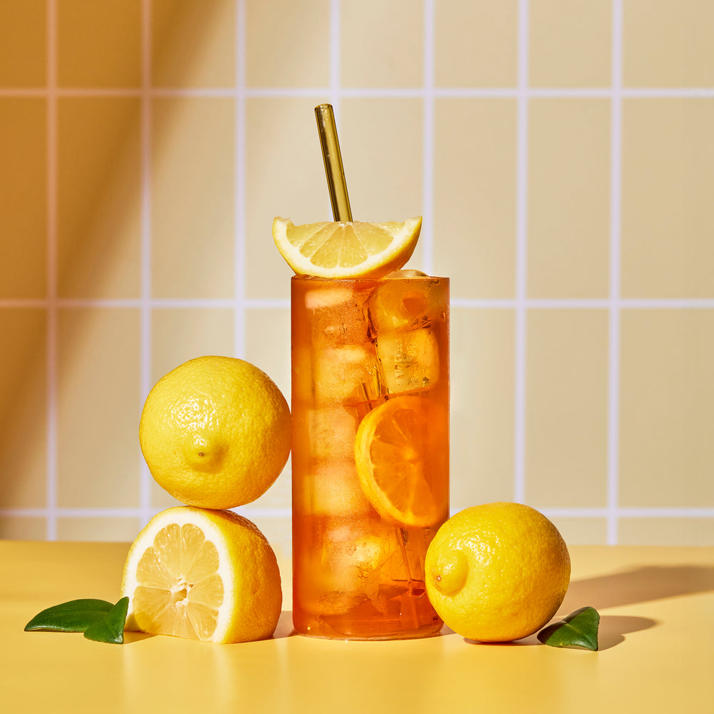 Full glass of Lemon Black iced tea with a lemon wedge garnish surrounded by whole and cut lemons on a yellow table.