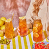 Several glasses of Lemon Black iced tea on a table with a bowl of tomatoes on the vine, a small bowl of green olives and a basket of lemons.