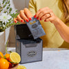 Person with red nails holding an overwrapped sachet of Lord Bergamot about to tear it open over a box of Lord Bergamot next to a bowl of citrus.