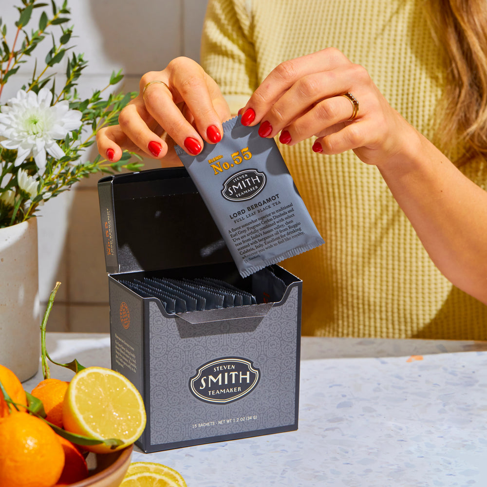 Person with red nails holding an overwrapped sachet of Lord Bergamot about to tear it open over a box of Lord Bergamot next to a bowl of citrus.