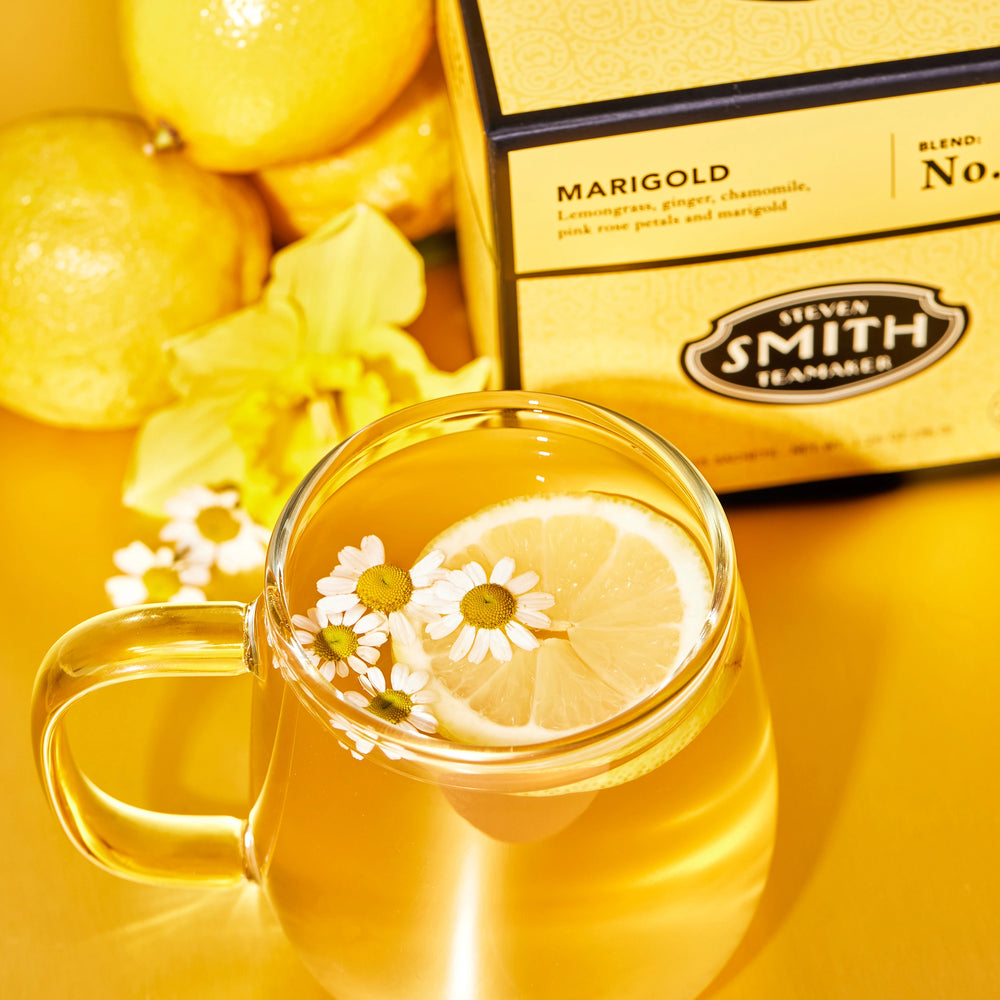 Brewed tea in glass with chamomile flowers and a lemon slice as garnish. Yellow Marigold package and lemons in background.