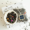 Small bowl of loose leaf Black Lavender tea next to a sachet of Black Lavender showing ingredient detail like full leaf black tea and lavender petals.