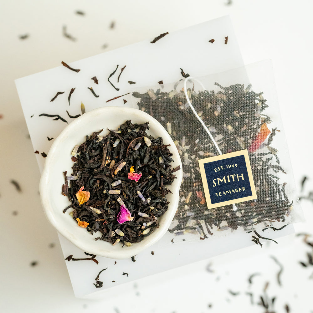 Small bowl of loose leaf Black Lavender tea next to a sachet of Black Lavender showing ingredient detail like full leaf black tea and lavender petals.