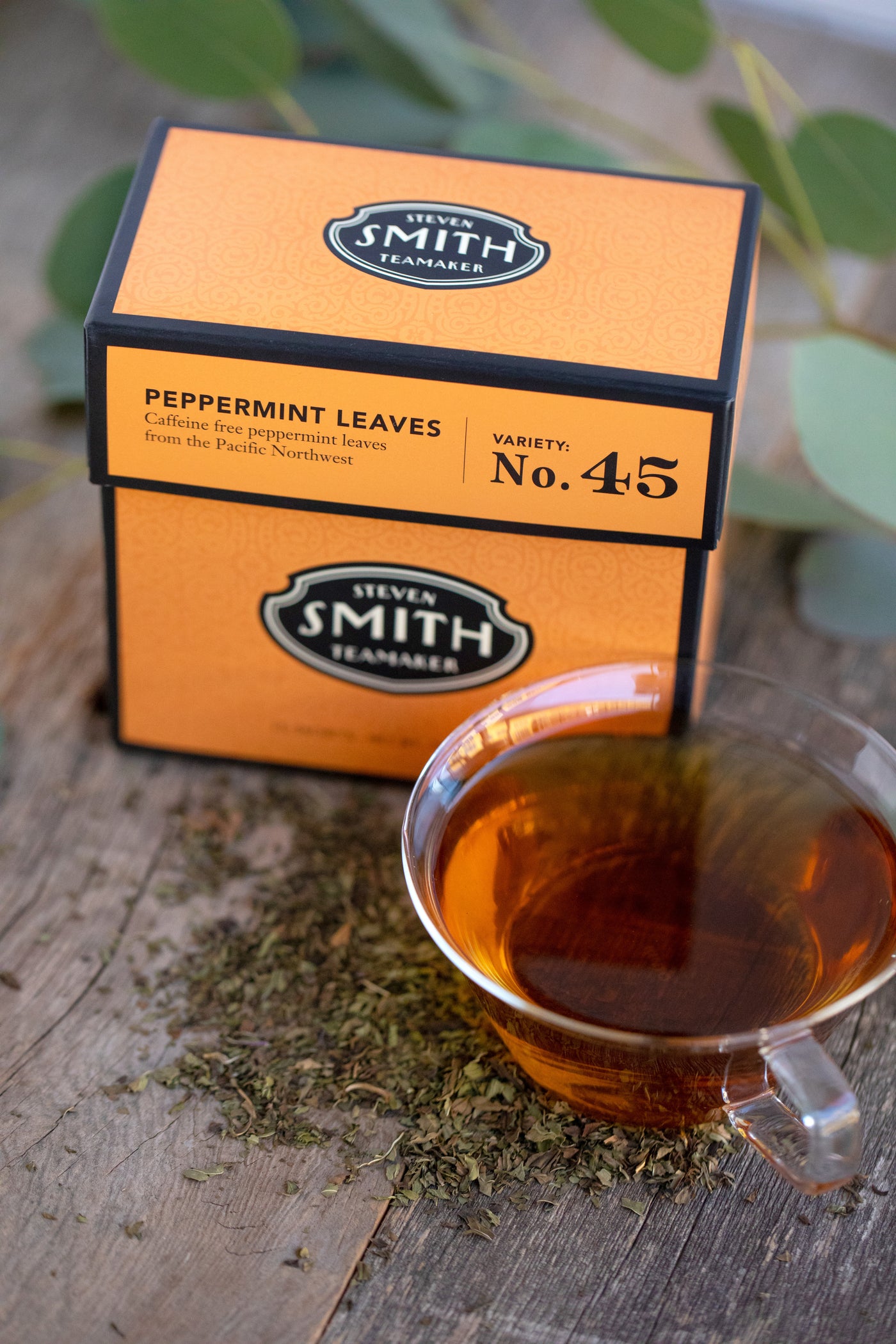 Orange box of Peppermint Leaves herbal infusion with loose peppermint leaves scattered on table next to a cup of brewed tea.