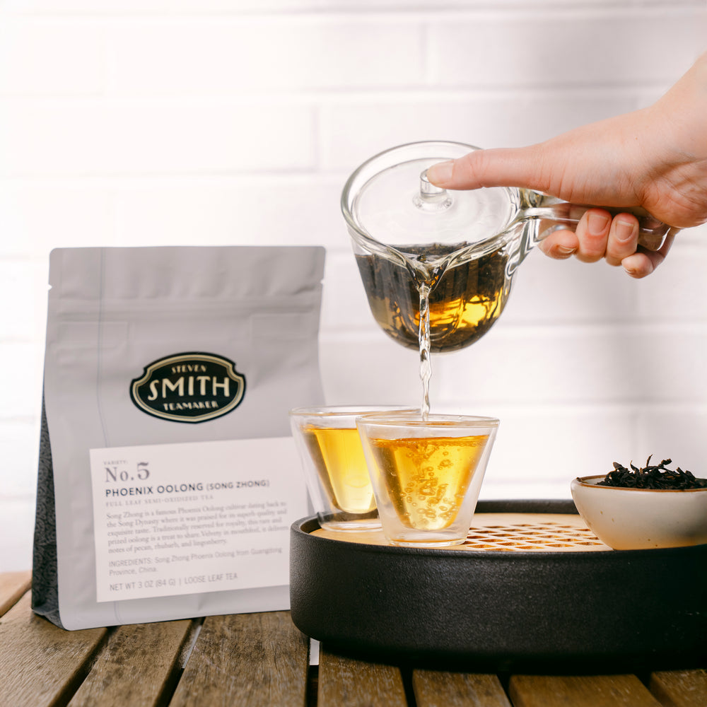 Loose leaf bag of Phoenix Oolong with brewed tea being poured into a small cup and a small dish with loose tea inside.