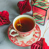 Ornate cream and red teacup and saucer filled with brewed tea with a carton of Santa's Little Helper in the background.