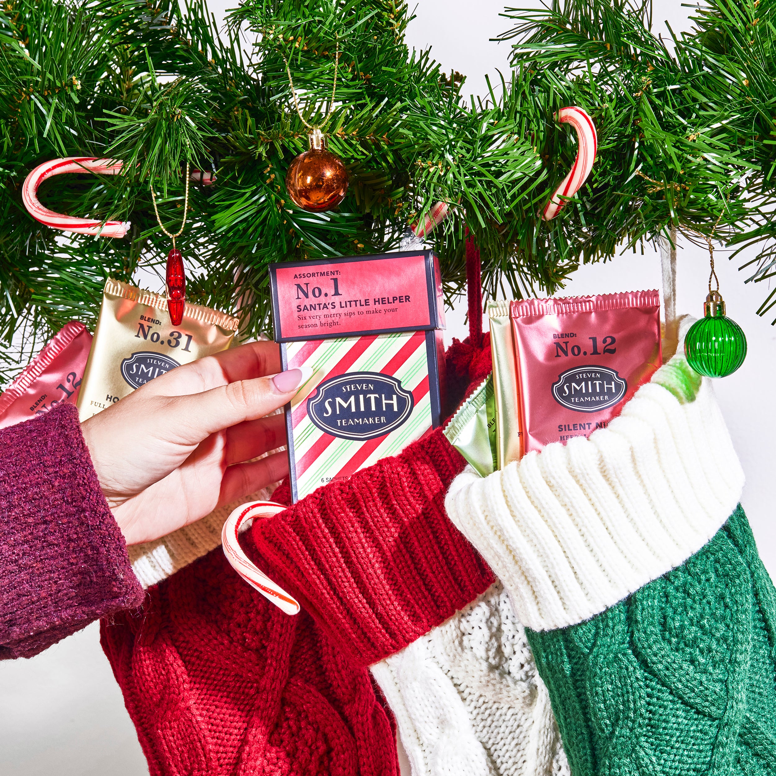Holiday scene with person placing Santa's Little Helper into a stocking next to two stockings filled with sachets of tea.
