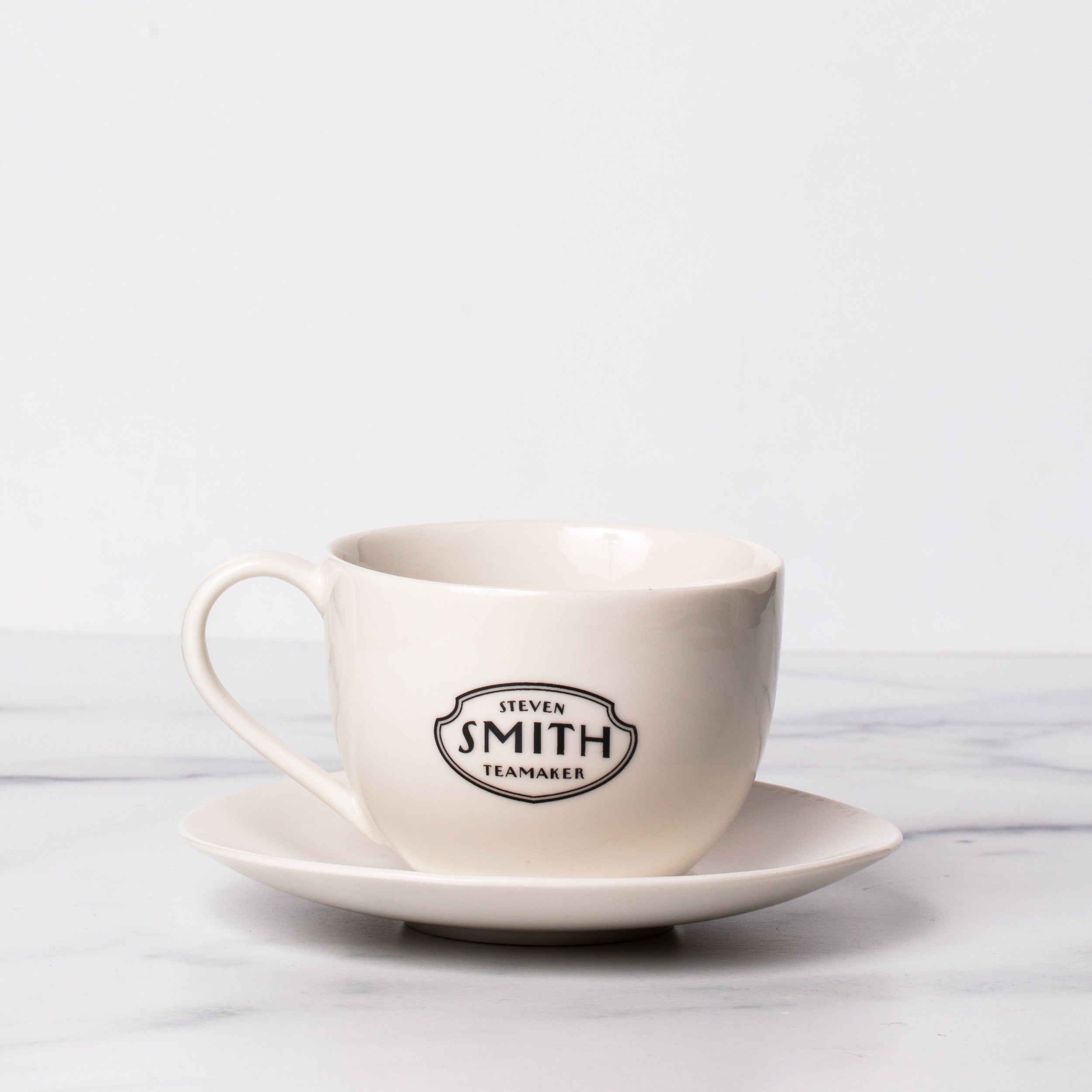 Porcelain teacup and saucer with black Smith logo shield on the front of teacup..