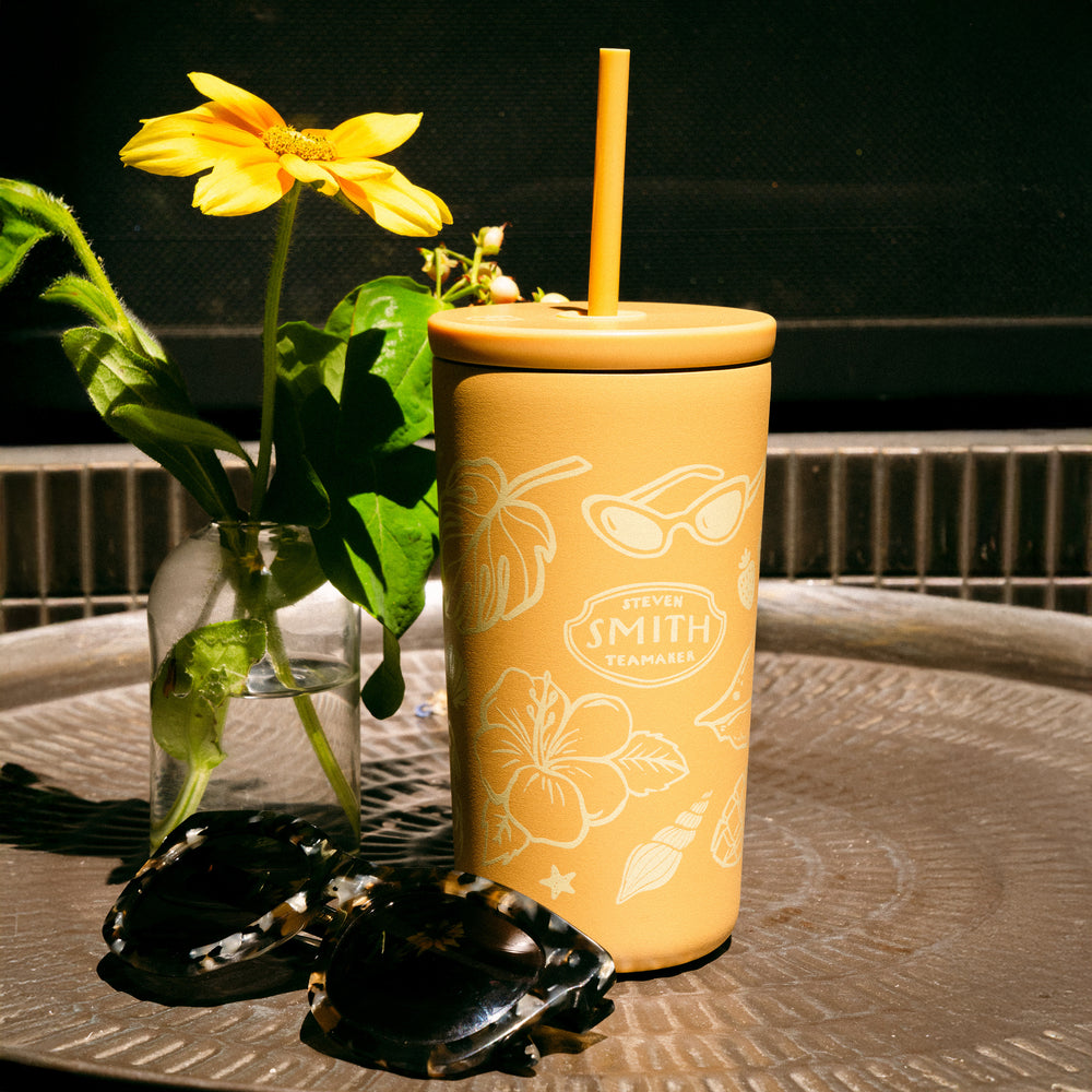 Yellow tumbler with floral design on a table with a vase of flowers and stones.