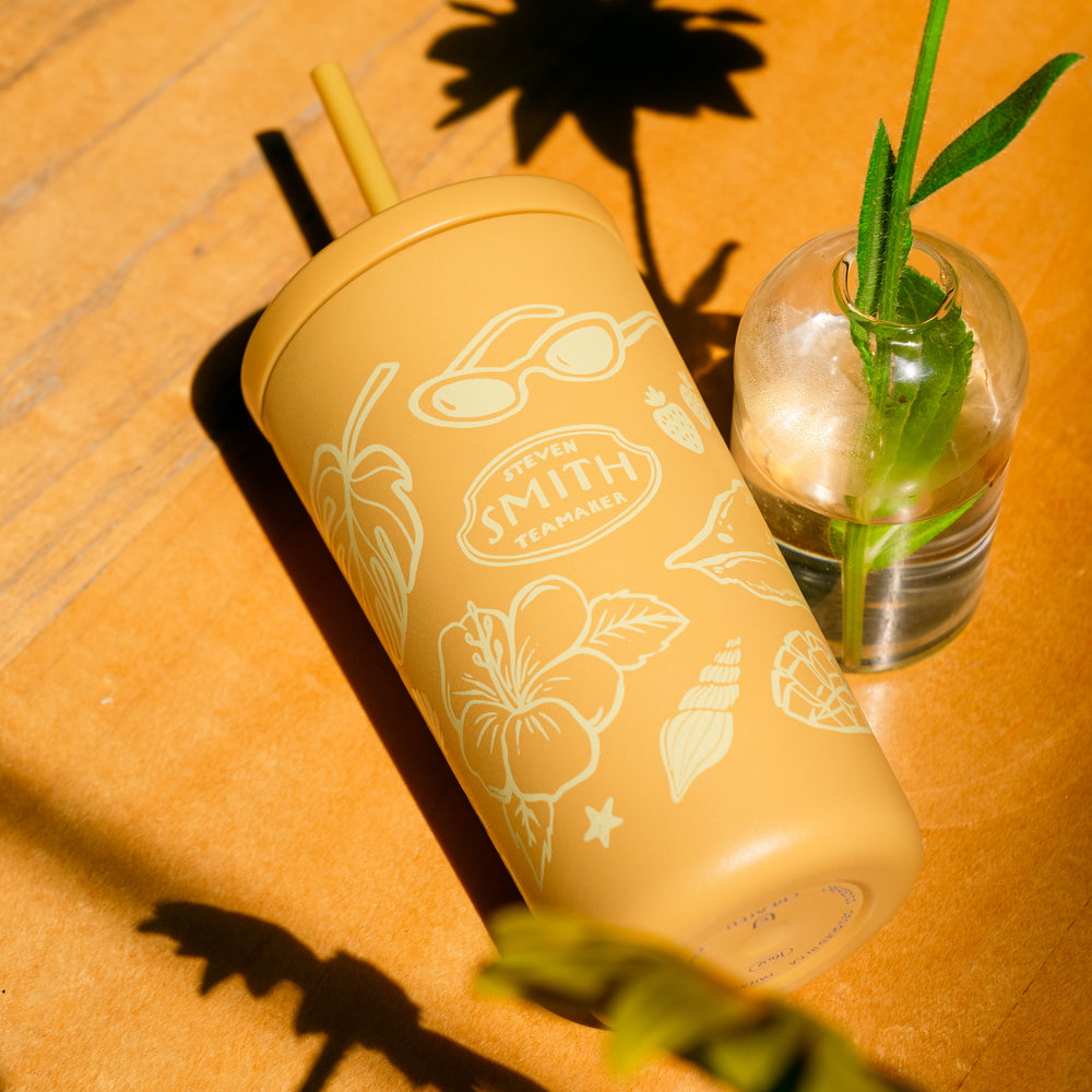 Yellow tumbler with floral design and straw on a wooden surface with a glass of water and plant.