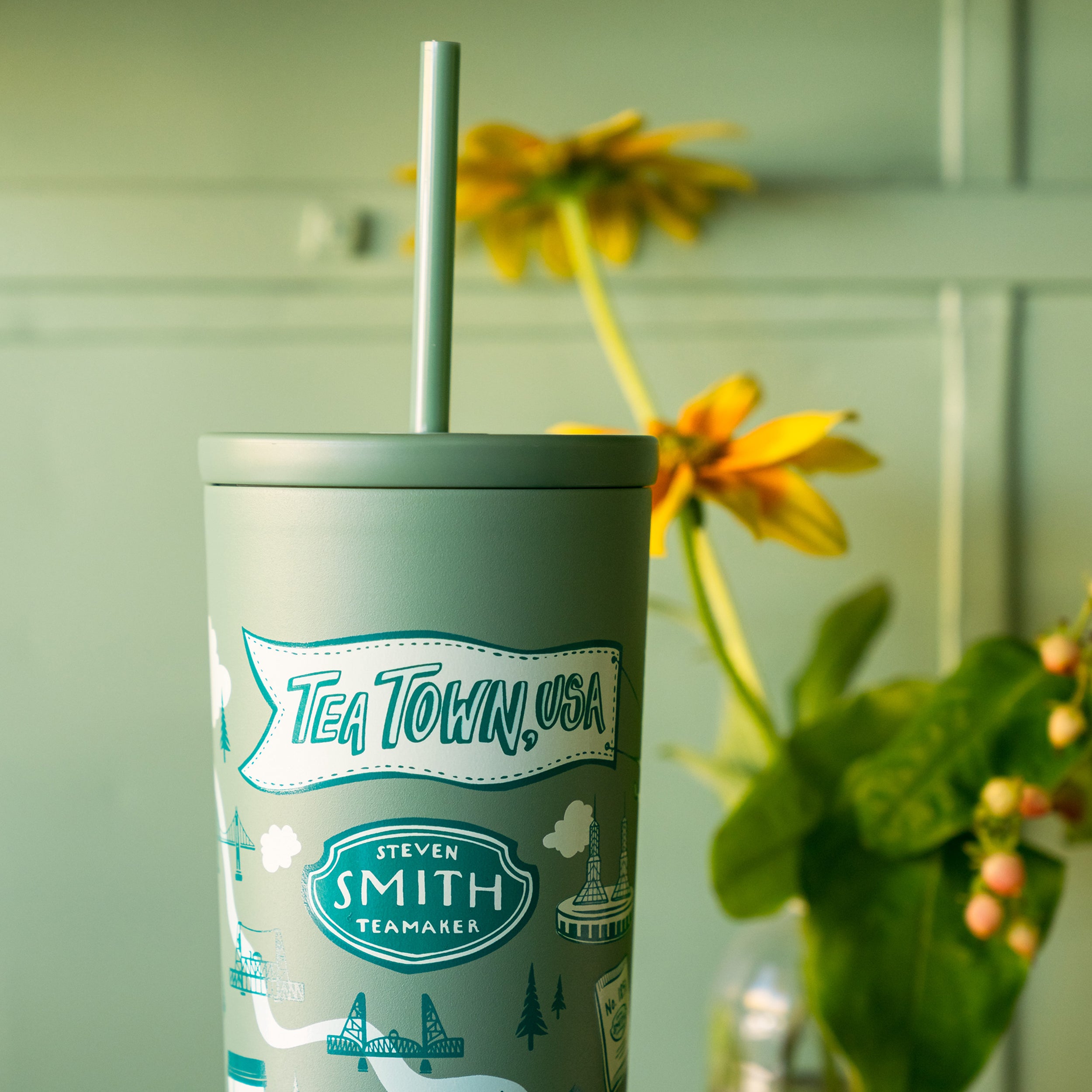 Green tumbler with 'Tea Town USA' branding and a straw, placed against a light green wall with flowers.