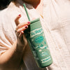 Person holding a green 'Tea Town' tumbler with a straw on a textured surface.