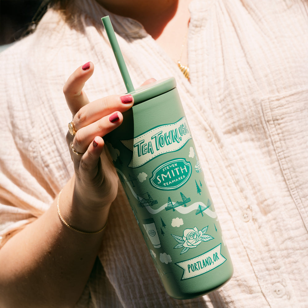 Person holding a green 'Tea Town' tumbler with a straw on a textured surface.