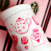 Small white beverage tumbler turned to show pink Valentine's Day design with teapot image, envelope with a love note and roses.