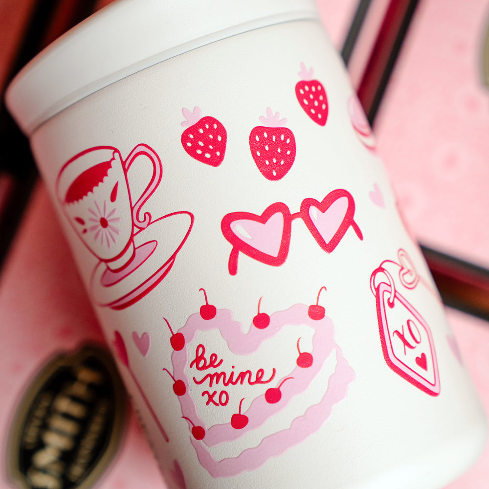 Small white beverage tumbler turned to show pink Valentine's Day design with teacup image, heart-eye glasses and a heart-shaped cake illustration.