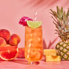 White Peach iced tea in glass with a lime, flower and straw on pink backdrop with peaches, orange and pineapple in background.