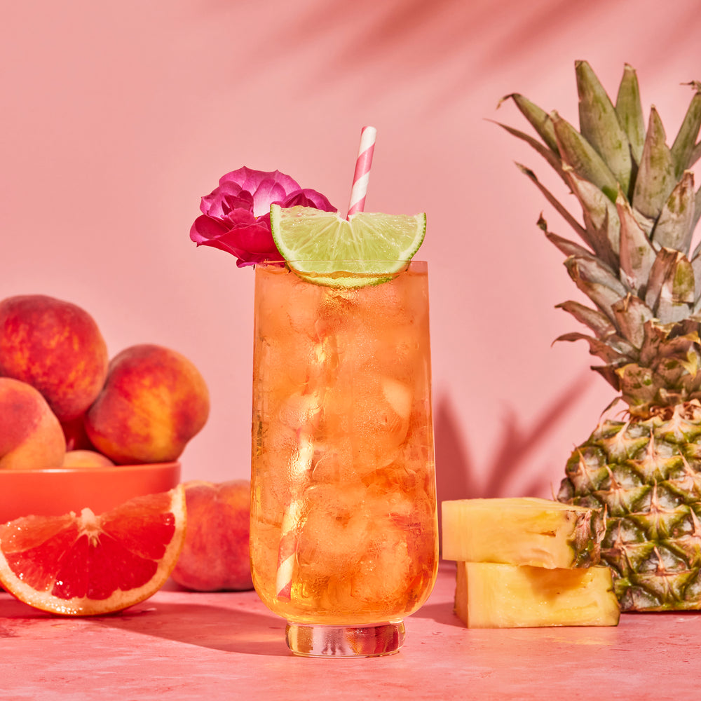 White Peach iced tea in glass with a lime, flower and straw on pink backdrop with peaches, orange and pineapple in background.